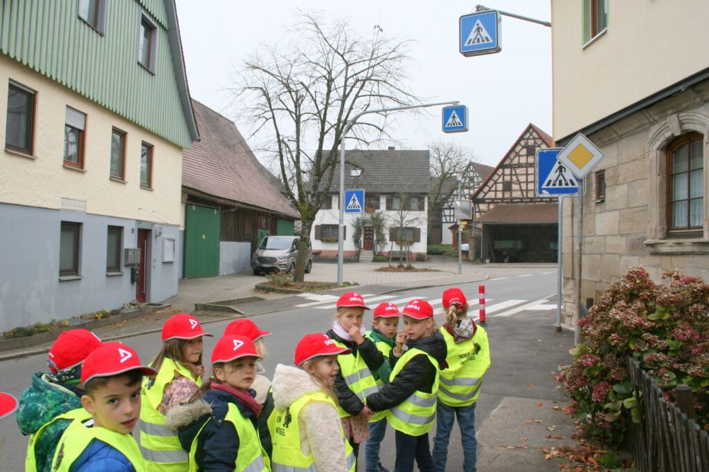 Erstklässler mit Warnweste und roter Schildmütze gehen auf einen Fußgängerüberweg zu. Fünf Verkehrsschilder sind zu sehen.