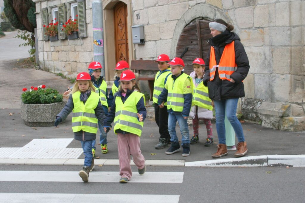 Zwei Erstklässlerinnen betreten gerade mit ausgestrecktem Arm den Zebrastreifen, fünf weitere warten mit einer Lehrerin auf dem Gehweg.