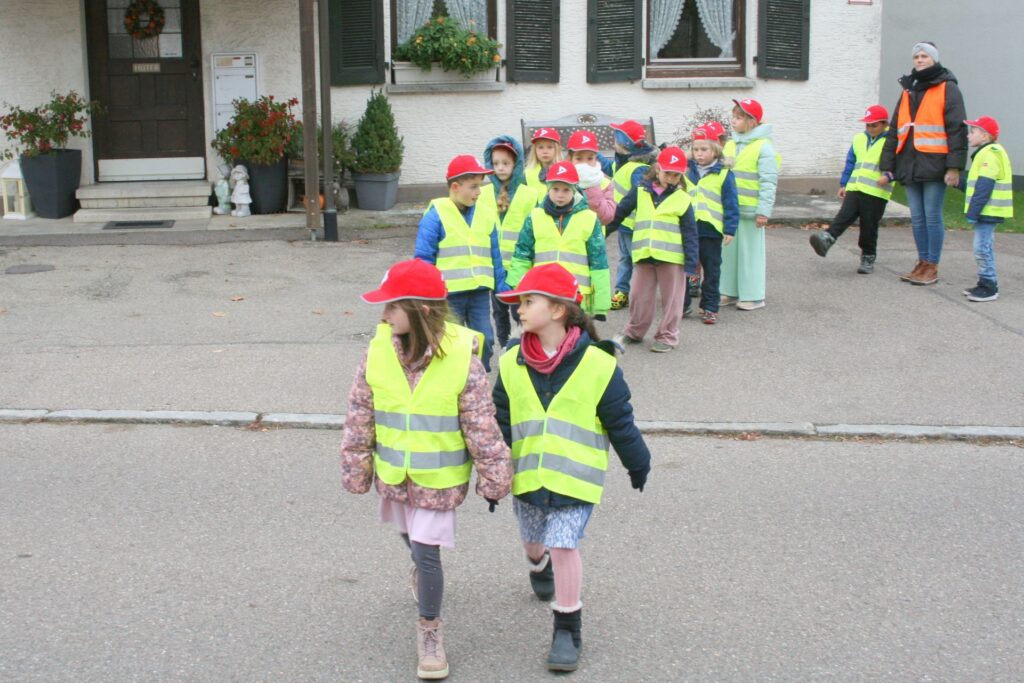 Zwei Erstklässlerinnen überqueren eine Straße ohne Überquerungshilfe. Auf dem Gehweg dahinter warten elf Klassenkameraden auf ihren Einsatz.