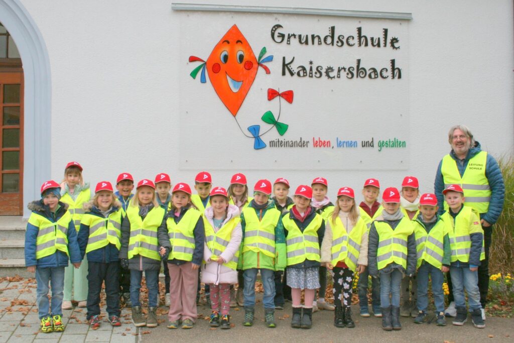 Klassenfoto aller 20 Kinder mit Verkehrswesten und roten Dekra-Mützen. Der Klassenlehrer steht hinten rechts. Er trägt auch eine Verkehrsweste.