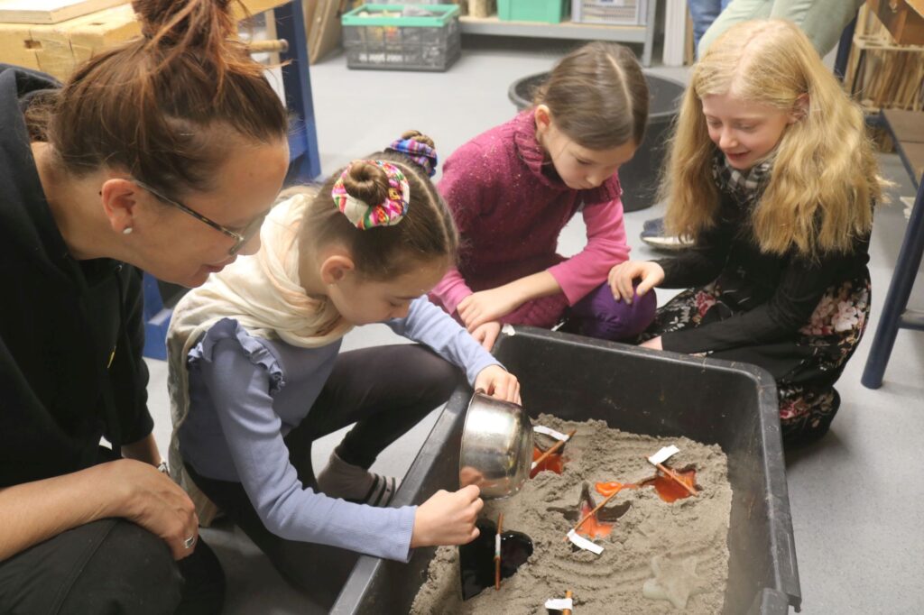 Kinder gießen Wachskerzen in einer Sandwanne.