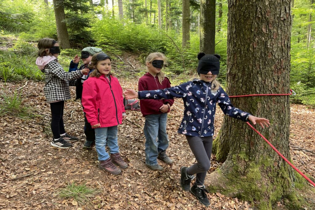 Vier Kinder mit verbundenen Augen hangeln sich am Seil des Blindenparcours im Wald entlang.